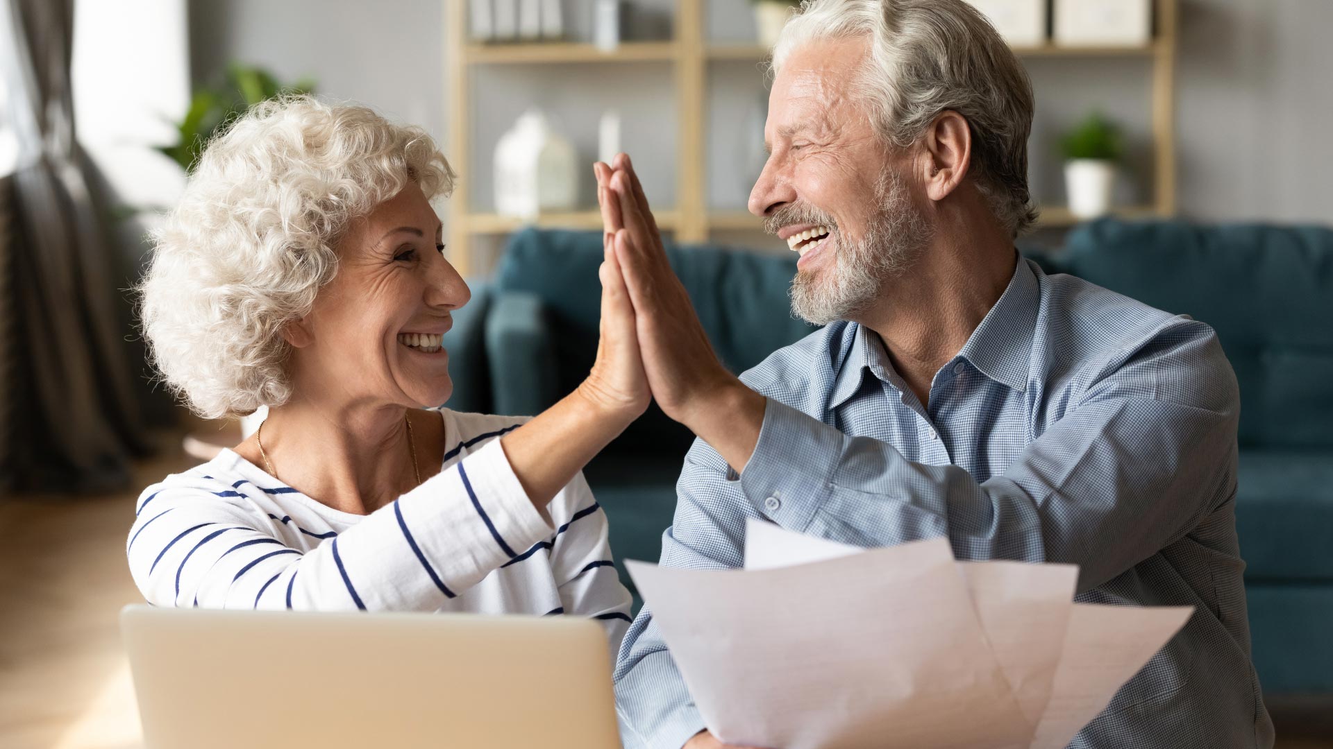 A happy elderly couple, a woman with curly white hair and a man with a grey beard, give each other a high-five. They both smile broadly, looking towards each other with closed eyes. The woman wears a white shirt with navy stripes, and a gold necklace. She is seated in front of a silver laptop to the left. The man wears a light blue checkered button-up shirt and holds several white papers in his left hand. The setting appears to be a cozy living room with a green couch in the blurred background.