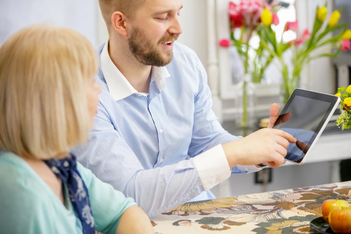 Two people sitting down at a table. One man is showing an older woman something on his tablet.